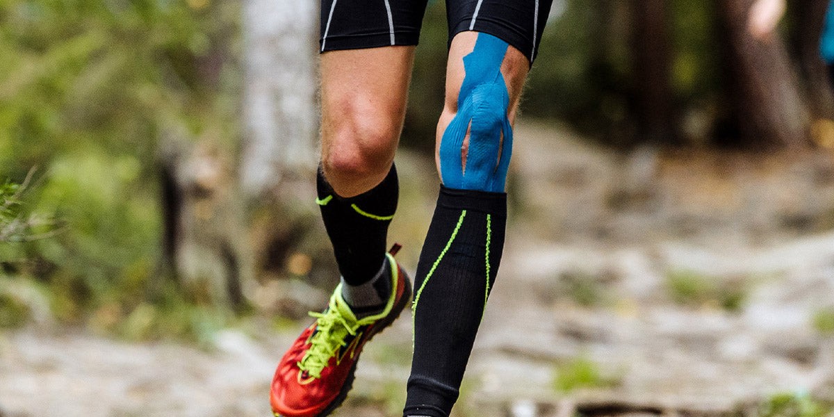 Trail runner's legs mid-stride with blue TapeGeeks kinesiology tape applied to the knee