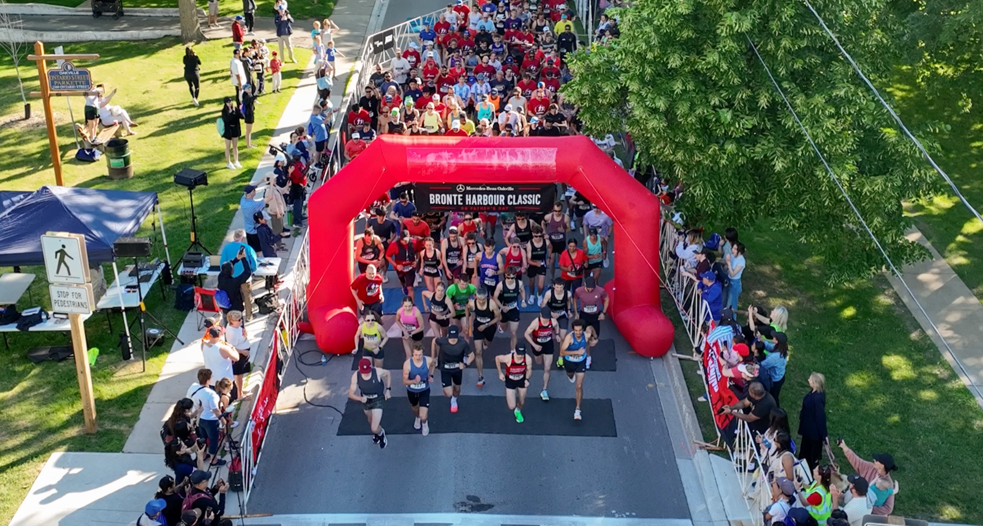 Aerial view of the densely packed start line at the Bronte Harbour Classic 5K, hundreds of runners streaming under the red Mercedes-Benz Oakville arch on a tree-lined Oakville street